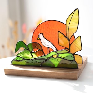 Op de afbeelding: Gebrandschilderde glazen sculptuur van een groene alligator met een gele oog, een witte vogel en een oranje zon. Het kunstwerk is gemonteerd op een houten voet, met gele en groene bladeren. Een uniek decoratief stuk.