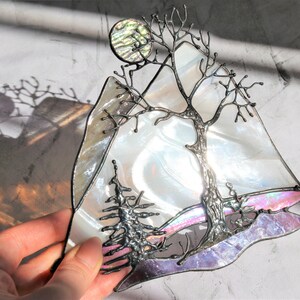 May include: A stained glass art piece depicting a tree with bare branches, a full moon, and a mountain range. The glass is iridescent and features shades of white, pink, and purple.