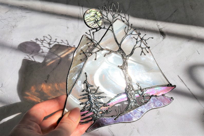 May include: A stained glass art piece depicting a tree with bare branches, a full moon, and a mountain range. The glass is iridescent and features shades of white, pink, and purple.