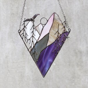May include: A stained glass window hanging with a chain. The window is shaped like a diamond and depicts a purple and pink mountain scene with a bird flying overhead.