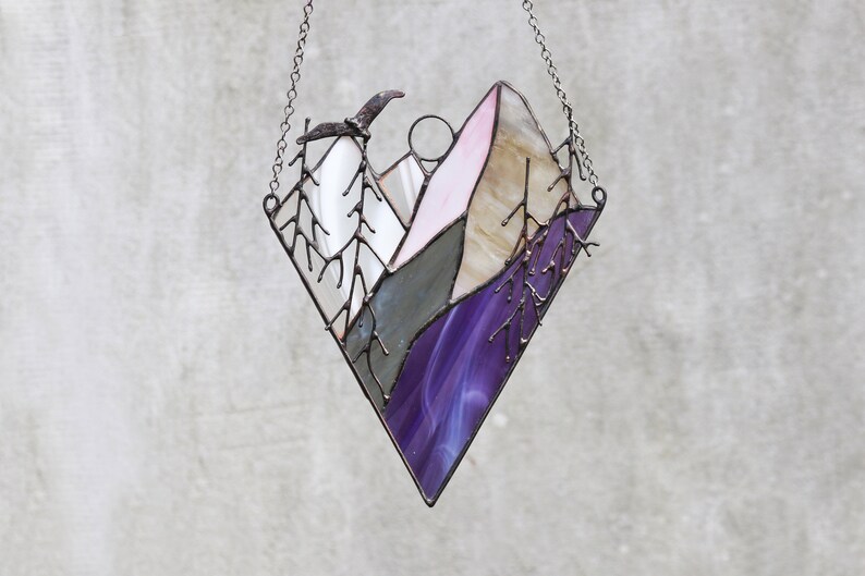 May include: A stained glass window hanging with a chain. The window is shaped like a diamond and depicts a purple and pink mountain scene with a bird flying overhead.