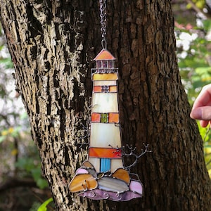 May include: A stained glass lighthouse ornament hanging from a chain on a tree trunk. The lighthouse is white with orange, red, and blue accents. The ornament is surrounded by a brown tree trunk.