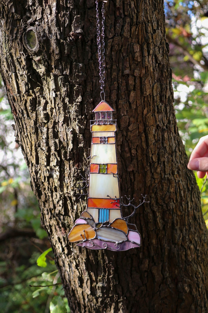 May include: A stained glass lighthouse ornament hanging from a chain on a tree trunk. The lighthouse is white with orange, red, and blue accents. The ornament is surrounded by a brown tree trunk.