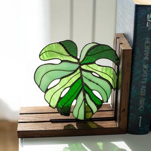 K&ouml;nnte beinhalten: Ein Buchst&uuml;tze in Form eines Monstera-Blattes aus Glasmalerei in Gr&uuml;nt&ouml;nen, mit einem dunkelbraunen Holzsockel. Das Blattdesign weist verschiedene Gr&uuml;nt&ouml;ne auf und ist vor einem wei&szlig;en Hintergrund platziert. Ein Buch befindet sich hinter der Buchst&uuml;tze.