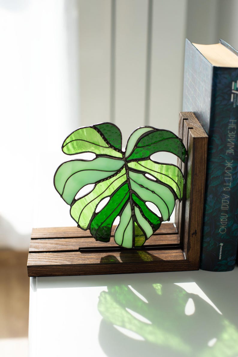 K&ouml;nnte beinhalten: Ein Buchst&uuml;tze in Form eines Monstera-Blattes aus Glasmalerei in Gr&uuml;nt&ouml;nen, mit einem dunkelbraunen Holzsockel. Das Blattdesign weist verschiedene Gr&uuml;nt&ouml;ne auf und ist vor einem wei&szlig;en Hintergrund platziert. Ein Buch befindet sich hinter der Buchst&uuml;tze.