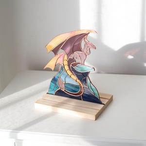 Op de afbeelding: Een gebrandschilderde drakenbeeldhouwwerk op een houten voet. De draak heeft een paars lichaam, gele vleugels en een blauwgroene basis. De sculptuur wordt tentoongesteld op een wit oppervlak.