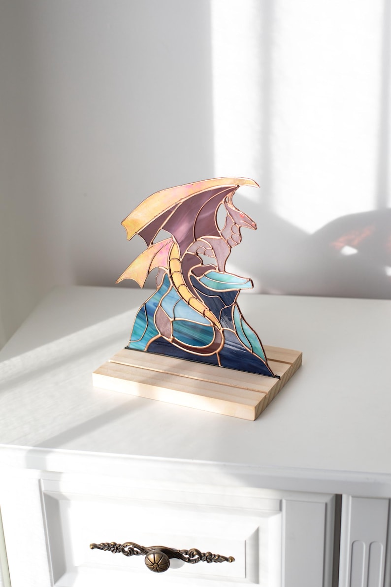 Op de afbeelding: Een gebrandschilderde drakenbeeldhouwwerk op een houten voet. De draak heeft een paars lichaam, gele vleugels en een blauwgroene basis. De sculptuur wordt tentoongesteld op een wit oppervlak.