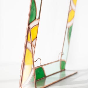 May include: A stained glass picture frame with a copper-colored metal frame. The frame features yellow, green, white, and light blue glass panels. The frame is standing upright on a clear glass base.