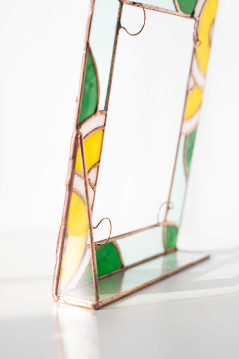 May include: A stained glass picture frame with a copper-colored metal frame. The frame features yellow, green, white, and light blue glass panels. The frame is standing upright on a clear glass base.