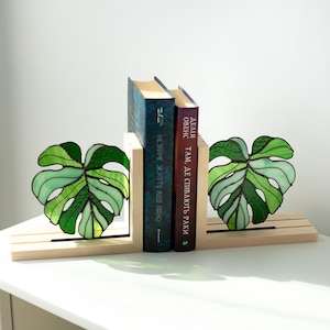 K&ouml;nnte beinhalten: Dekorative Buchst&uuml;tzen mit einem Design aus Glasmalerei in Form eines Monstera-Blattes in Gr&uuml;nt&ouml;nen. Die Buchst&uuml;tzen aus Holz halten zwei B&uuml;cher. Die Buchst&uuml;tzen stehen auf einer wei&szlig;en Oberfl&auml;che.
