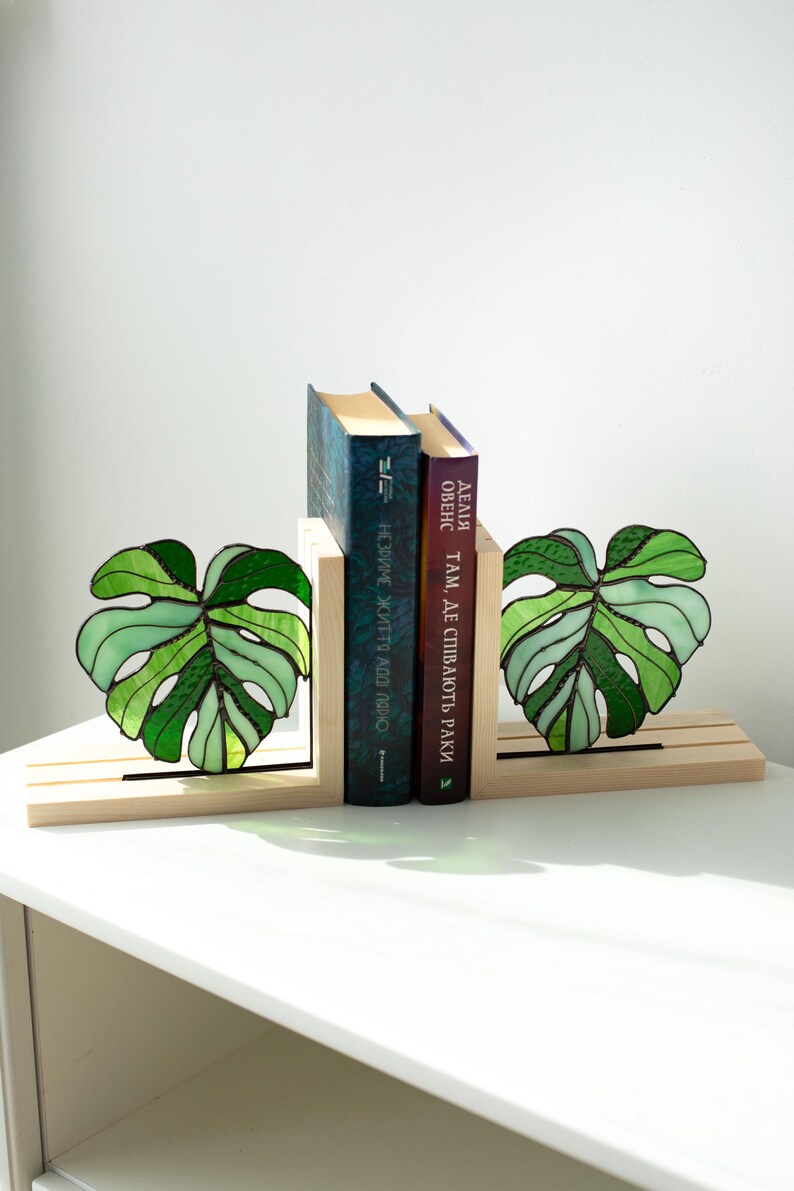 K&ouml;nnte beinhalten: Dekorative Buchst&uuml;tzen mit einem Design aus Glasmalerei in Form eines Monstera-Blattes in Gr&uuml;nt&ouml;nen. Die Buchst&uuml;tzen aus Holz halten zwei B&uuml;cher. Die Buchst&uuml;tzen stehen auf einer wei&szlig;en Oberfl&auml;che.