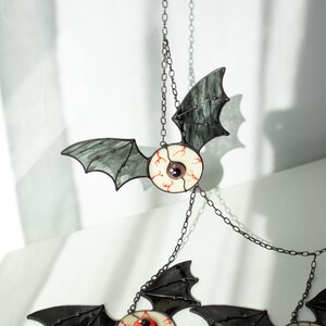 May include: A hanging mobile featuring three stained glass bat ornaments. Each bat has dark grey translucent wings and a central circular eye with a white sclera, red veins, and a coloured iris in purple, orange, or blue, connected by metal chains.