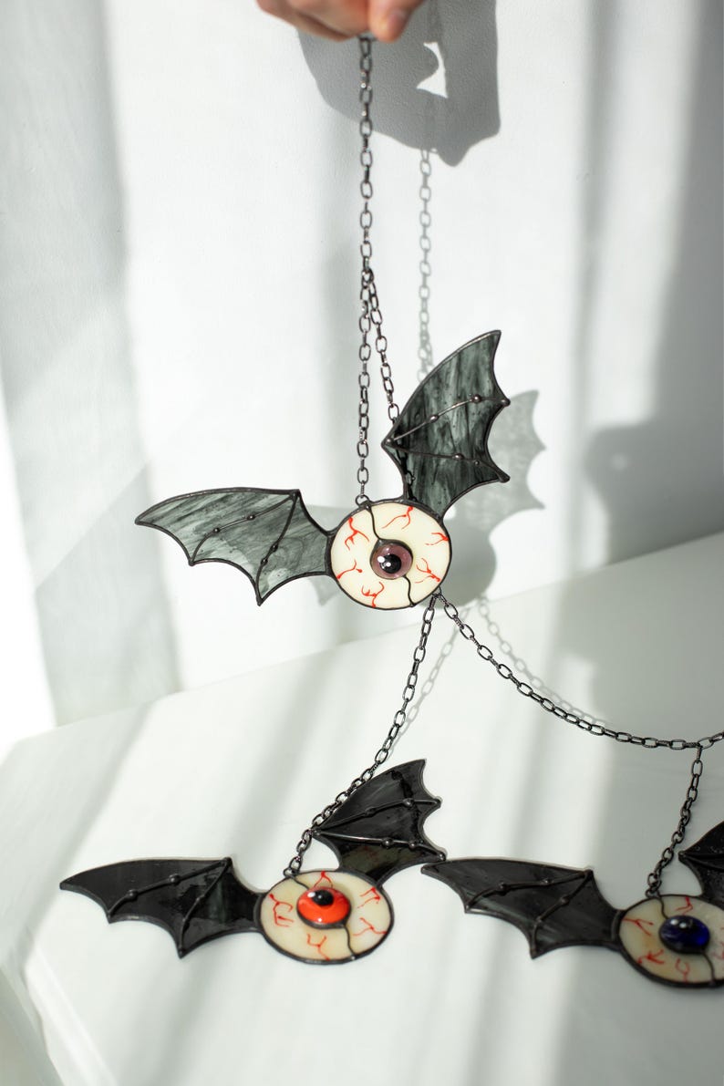 May include: A hanging mobile featuring three stained glass bat ornaments. Each bat has dark grey translucent wings and a central circular eye with a white sclera, red veins, and a coloured iris in purple, orange, or blue, connected by metal chains.