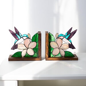 Pu&ograve; includere: Un paio di fermalibri in legno con un design a vetrata. Ogni fermalibro raffigura un colibr&igrave; con ali turchesi e viola, vicino a un fiore rosa e foglie verdi. I fermalibri hanno una base in legno marrone scuro.