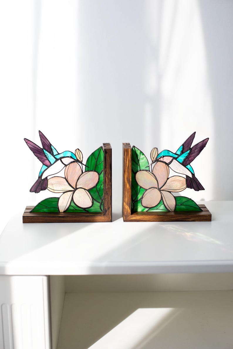 Pu&ograve; includere: Un paio di fermalibri in legno con un design a vetrata. Ogni fermalibro raffigura un colibr&igrave; con ali turchesi e viola, vicino a un fiore rosa e foglie verdi. I fermalibri hanno una base in legno marrone scuro.