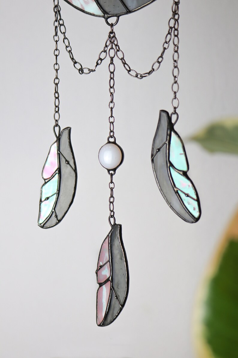 May include: A stained glass dreamcatcher with three feathers and a pearl bead hanging from a chain. The feathers are gray with pink and blue accents.