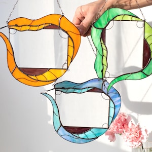 May include: Three stained glass snake-shaped frames in orange, green, and blue. Each frame is suspended by a chain and features a rectangular opening. The frames are displayed against a white background with pink flowers.