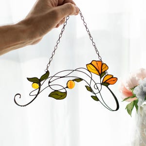 May include: A stained glass suncatcher featuring orange flowers and yellow berries with green leaves, connected by dark metal wire stems and attached to a silver-toned metal chain for hanging, showcasing translucent glass textures.