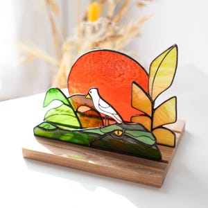 Op de afbeelding: Gebrandschilderd glaskunstwerk met een groene alligator met een gele oog, een witte vogel en een oranje zon. Het kunstwerk staat op een houten voet, met gele en groene bladeren. Een decoratief object.
