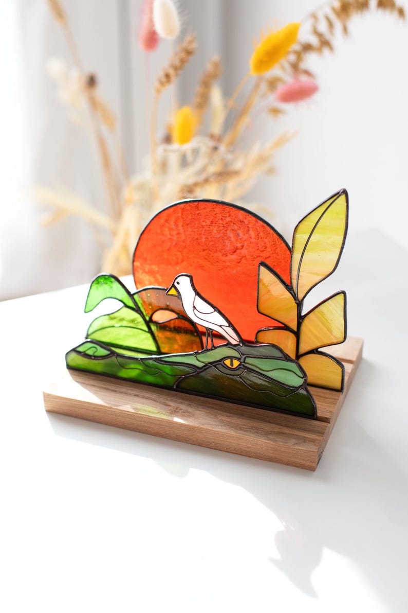 Op de afbeelding: Gebrandschilderd glaskunstwerk met een groene alligator met een gele oog, een witte vogel en een oranje zon. Het kunstwerk staat op een houten voet, met gele en groene bladeren. Een decoratief object.
