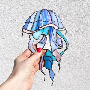 Op de afbeelding: Een glas-in-lood kwal met iriserende blauwe, paarse en roze glasstukken. De kwal heeft een klokvormige bovenkant en lange, vloeiende tentakels. De tentakels zijn versierd met kleine, heldere glazen kralen.