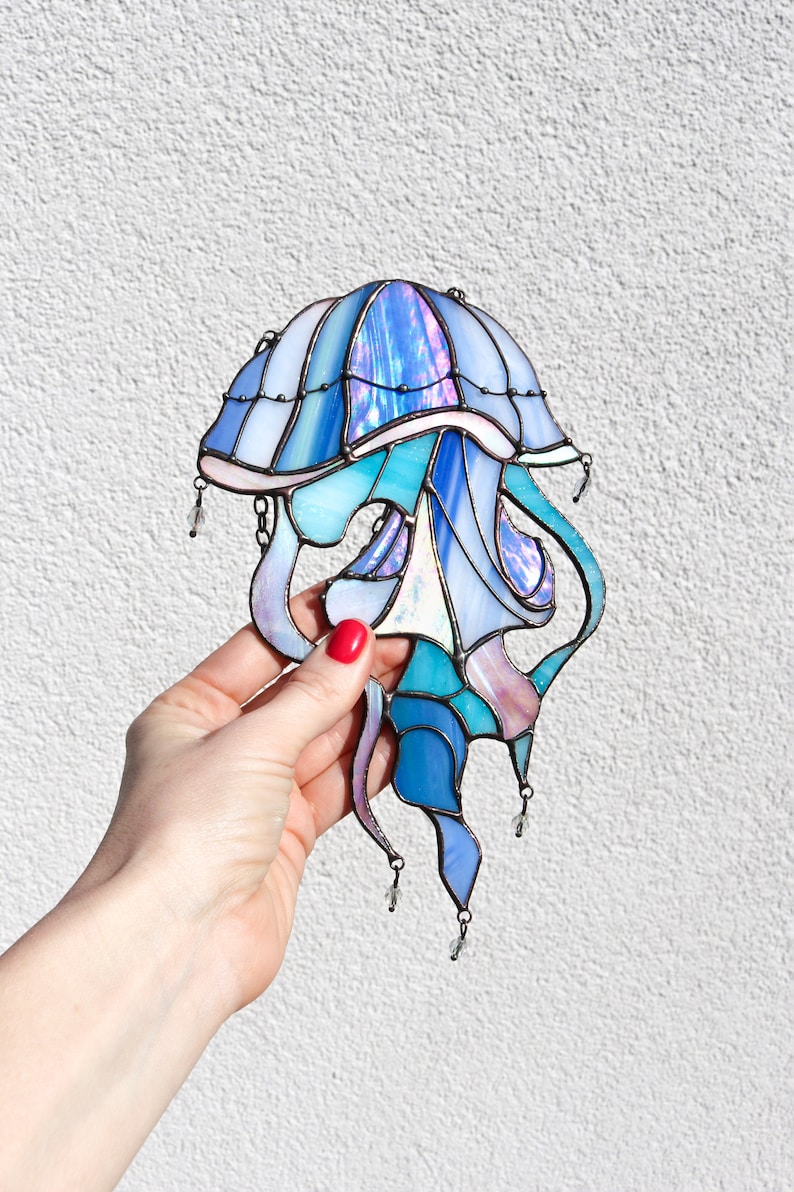 Op de afbeelding: Een glas-in-lood kwal met iriserende blauwe, paarse en roze glasstukken. De kwal heeft een klokvormige bovenkant en lange, vloeiende tentakels. De tentakels zijn versierd met kleine, heldere glazen kralen.