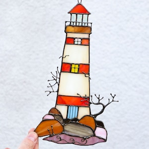 May include: A stained glass window hanging with a chain, depicting a red and white lighthouse on a rocky shore with brown and pink rocks. The lighthouse has a white base with red stripes and a small blue door.