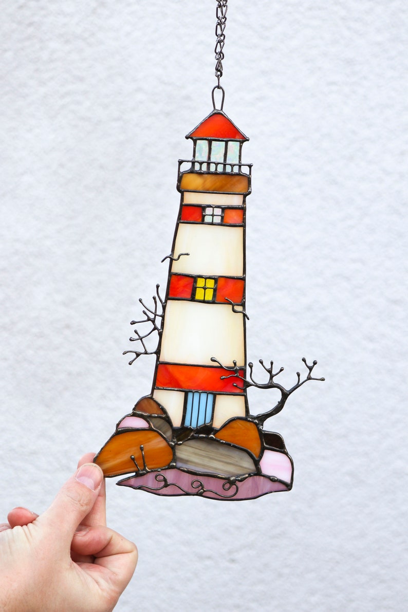 May include: A stained glass window hanging with a chain, depicting a red and white lighthouse on a rocky shore with brown and pink rocks. The lighthouse has a white base with red stripes and a small blue door.