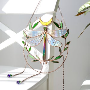 May include: A stained glass dragonfly suncatcher with iridescent wings, green leaves, and a yellow crescent moon. The suncatcher hangs from a copper chain with purple glass beads.