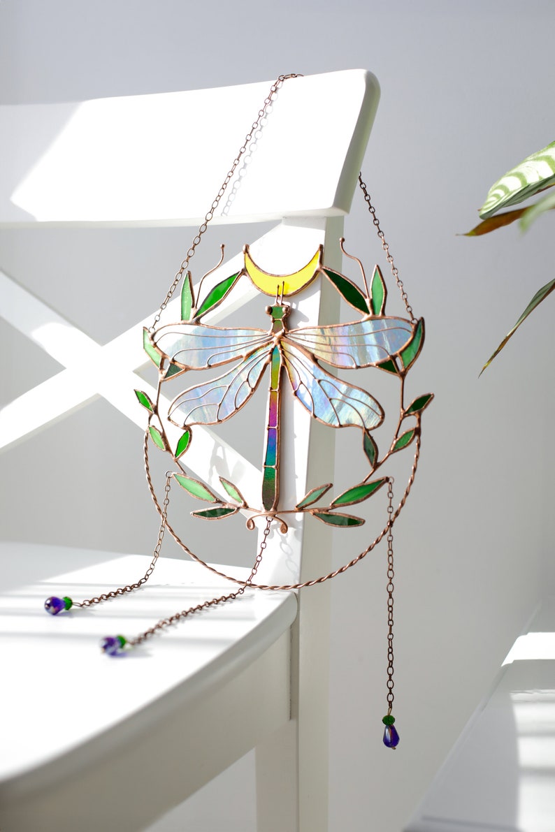 May include: A stained glass dragonfly suncatcher with iridescent wings, green leaves, and a yellow crescent moon. The suncatcher hangs from a copper chain with purple glass beads.