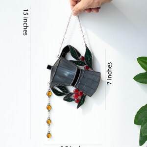 Puede incluir: Un atrapasoles de vidrieras con forma de cafetera moka, con vidrio gris oscuro, hojas verdes y bayas de caf&eacute; rojas. Una cadena de cuentas de vidrio color &aacute;mbar cuelga del pico. La pieza mide 17,78 cm de alto y 16,51 cm de ancho, con 38,1 cm de largo.