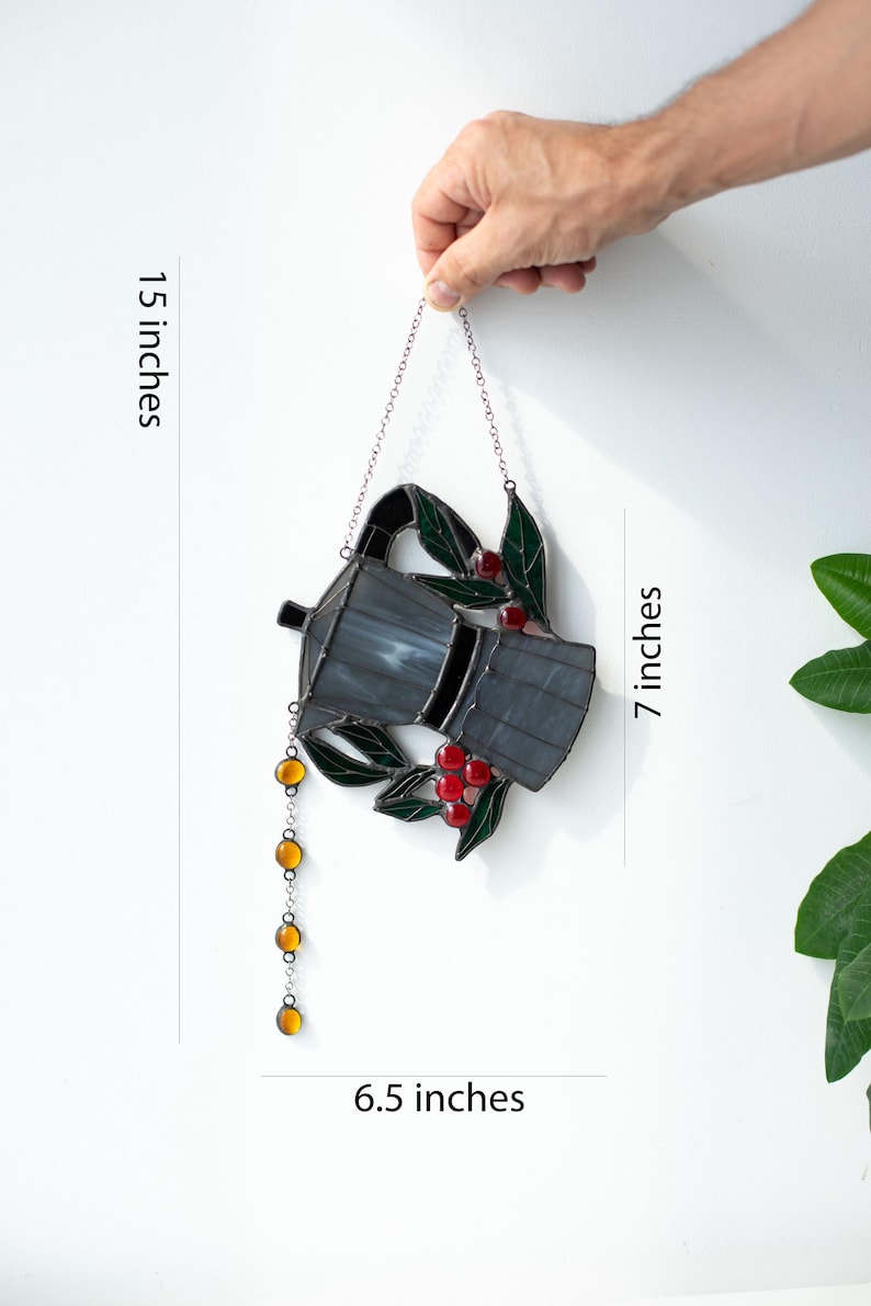 Puede incluir: Un atrapasoles de vidrieras con forma de cafetera moka, con vidrio gris oscuro, hojas verdes y bayas de caf&eacute; rojas. Una cadena de cuentas de vidrio color &aacute;mbar cuelga del pico. La pieza mide 17,78 cm de alto y 16,51 cm de ancho, con 38,1 cm de largo.