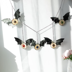 May include: A hanging mobile featuring five stained glass bats with central eyeballs. Each bat has black wings and a round eye with red veins, featuring irises in various colours including brown, green, red, blue, and hazel, connected by chains.