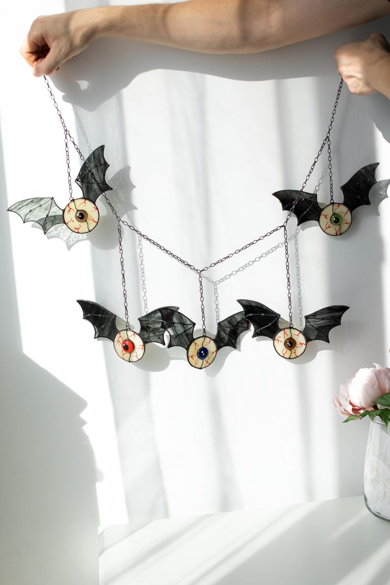 May include: A hanging mobile featuring five stained glass bats with central eyeballs. Each bat has black wings and a round eye with red veins, featuring irises in various colours including brown, green, red, blue, and hazel, connected by chains.