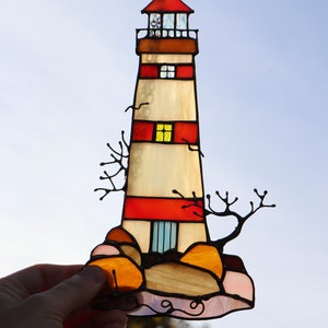 May include: A stained glass lighthouse with red and white stripes, a yellow window, and a blue door. The lighthouse is on a rocky base with orange and pink rocks. The stained glass is set against a blue sky.