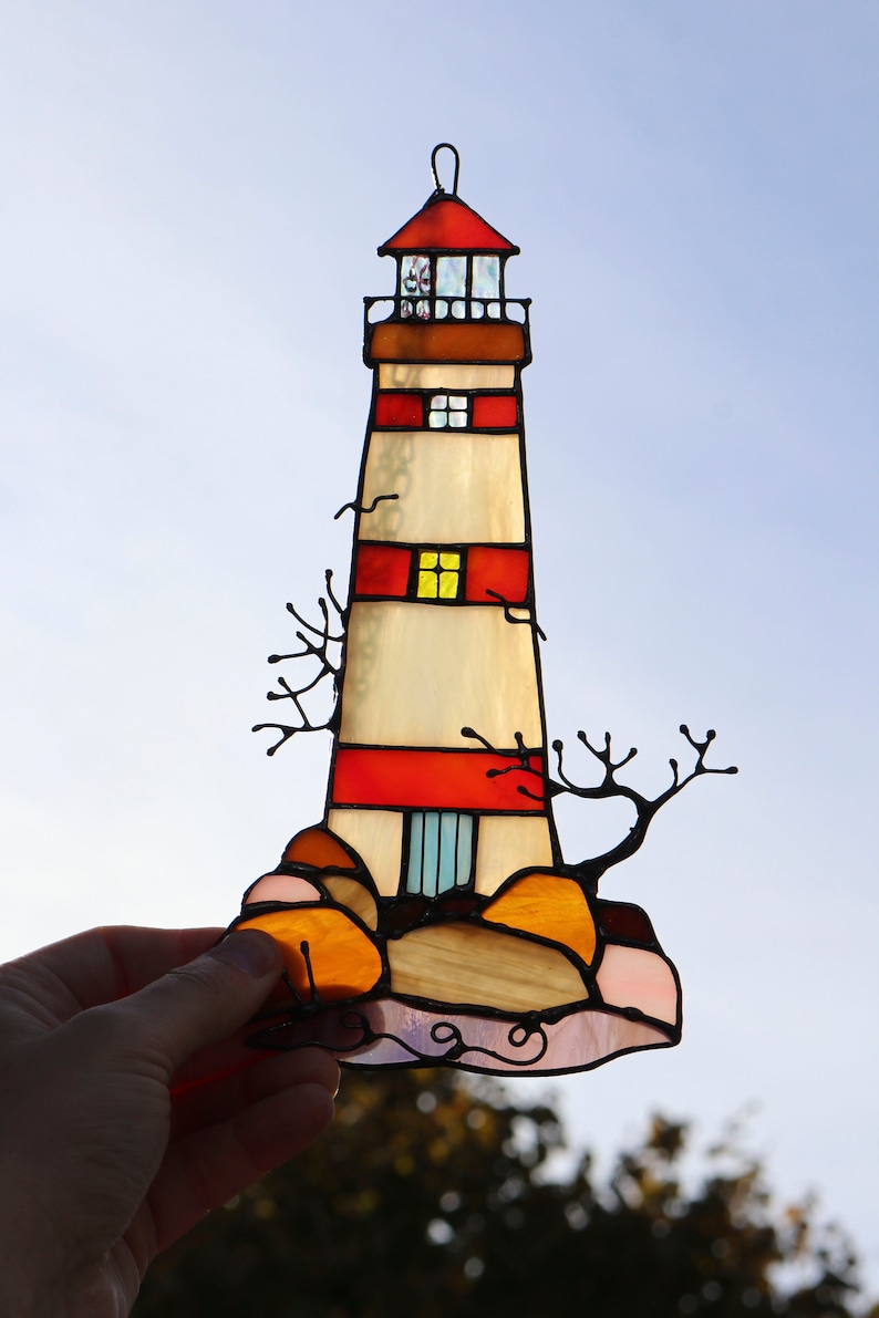 May include: A stained glass lighthouse with red and white stripes, a yellow window, and a blue door. The lighthouse is on a rocky base with orange and pink rocks. The stained glass is set against a blue sky.