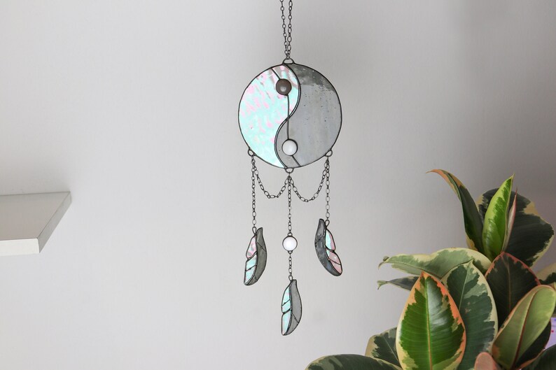 May include: A stained glass yin yang symbol with iridescent blue and white colors hangs from a chain with three iridescent blue and white feathers.