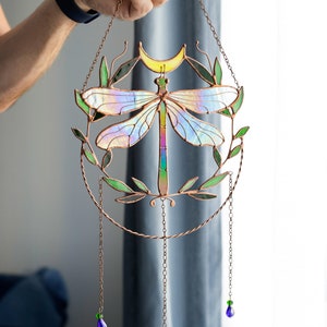 May include: A stained glass dragonfly suncatcher with iridescent wings and a copper frame. The dragonfly is surrounded by green leaves and a crescent moon. The suncatcher hangs from a copper chain with blue and green glass beads.