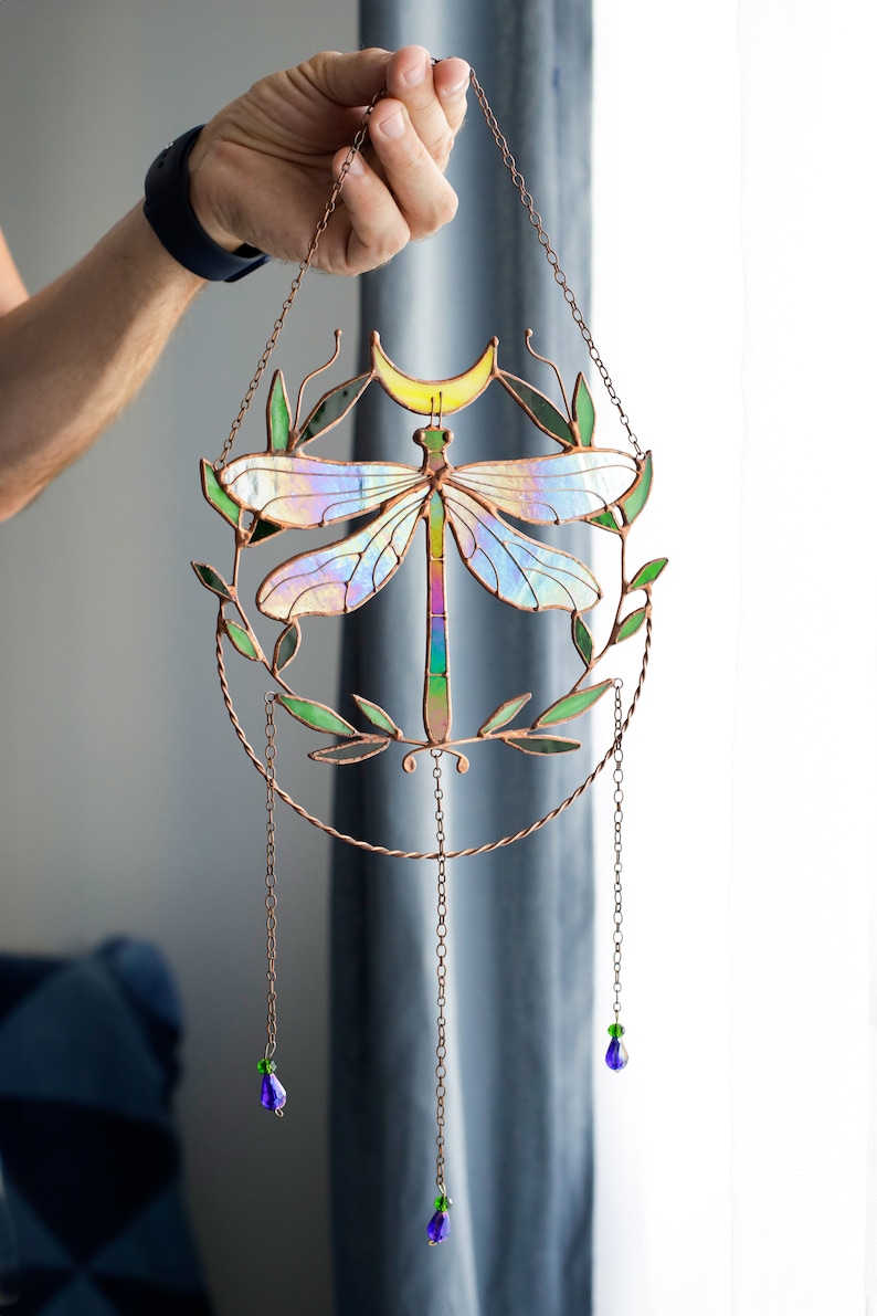 May include: A stained glass dragonfly suncatcher with iridescent wings and a copper frame. The dragonfly is surrounded by green leaves and a crescent moon. The suncatcher hangs from a copper chain with blue and green glass beads.