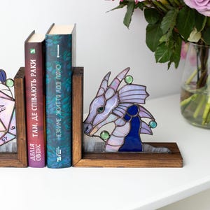 May include: A stained glass dragon bookend in shades of purple, blue, and green, with a wooden base. The dragon is positioned next to two books with Ukrainian text. A vase of purple roses is in the background.