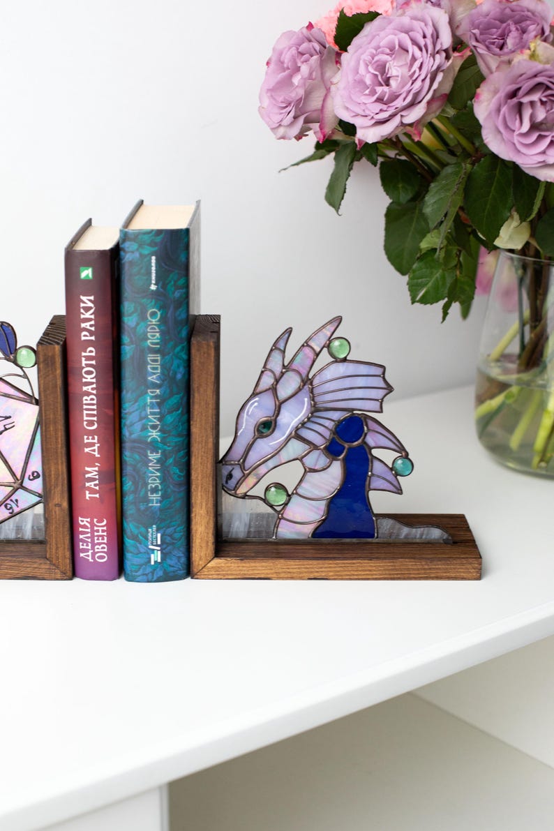 May include: A stained glass dragon bookend in shades of purple, blue, and green, with a wooden base. The dragon is positioned next to two books with Ukrainian text. A vase of purple roses is in the background.