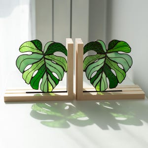 K&ouml;nnte beinhalten: Zwei Buchst&uuml;tzen mit Monstera-Blatt-Motiv aus Glasmalerei. Die Bl&auml;tter sind in verschiedenen Gr&uuml;nt&ouml;nen mit schwarzen Umrissen gehalten und stehen auf hellen Holzsockeln. Die Buchst&uuml;tzen stehen auf einer wei&szlig;en Oberfl&auml;che.