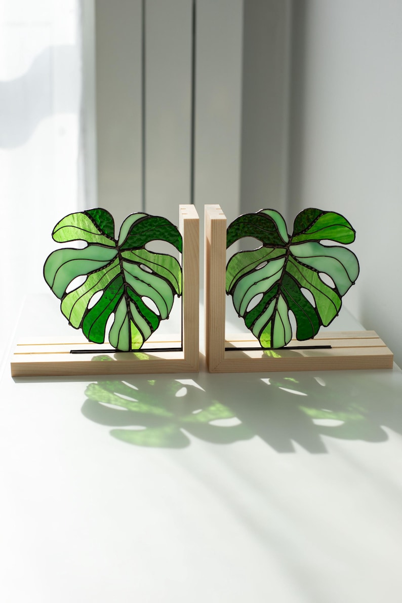 K&ouml;nnte beinhalten: Zwei Buchst&uuml;tzen mit Monstera-Blatt-Motiv aus Glasmalerei. Die Bl&auml;tter sind in verschiedenen Gr&uuml;nt&ouml;nen mit schwarzen Umrissen gehalten und stehen auf hellen Holzsockeln. Die Buchst&uuml;tzen stehen auf einer wei&szlig;en Oberfl&auml;che.