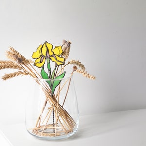 May include: A yellow stained glass flower with green leaves in a clear glass vase with dried wheat stalks.