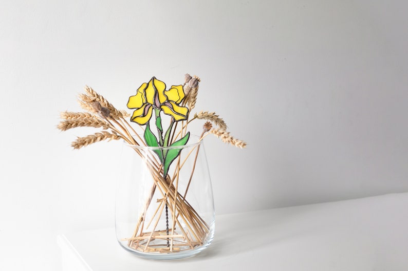 May include: A yellow stained glass flower with green leaves in a clear glass vase with dried wheat stalks.