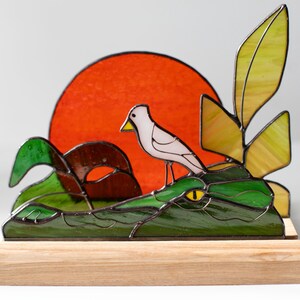 Op de afbeelding: Gebrandschilderde glazen sculptuur van een vogel op een groene alligator, met een rode zon. De sculptuur is gemonteerd op een houten voet. De vogel is wit met een gele snavel.