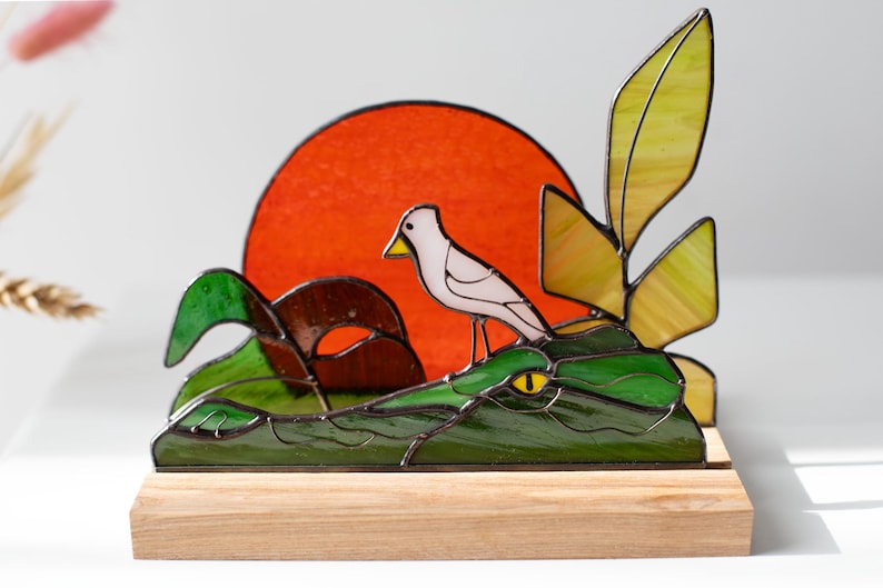 Op de afbeelding: Gebrandschilderde glazen sculptuur van een vogel op een groene alligator, met een rode zon. De sculptuur is gemonteerd op een houten voet. De vogel is wit met een gele snavel.