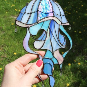 Op de afbeelding: Een glas-in-lood kwal met iriserende blauwe, roze en witte glas. De kwal heeft een klokvormige bovenkant en lange, vloeiende tentakels. Het glas wordt in een hand gehouden tegen een groene gras achtergrond.
