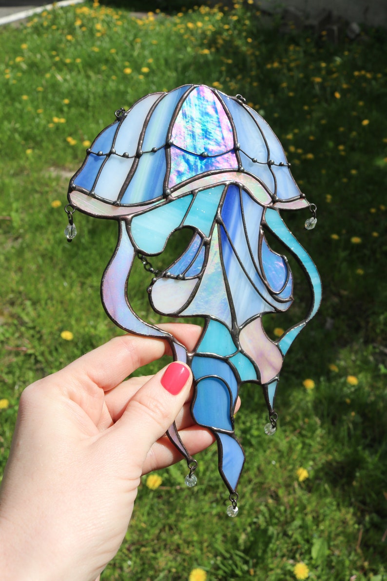 Op de afbeelding: Een glas-in-lood kwal met iriserende blauwe, roze en witte glas. De kwal heeft een klokvormige bovenkant en lange, vloeiende tentakels. Het glas wordt in een hand gehouden tegen een groene gras achtergrond.