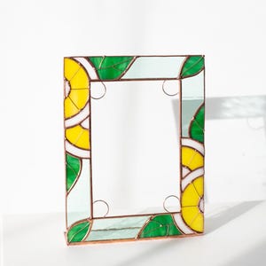 May include: A rectangular stained glass mirror with a vibrant design. The frame features yellow, green, and white geometric patterns, resembling lemons and leaves. The mirror is supported by a copper-colored base.